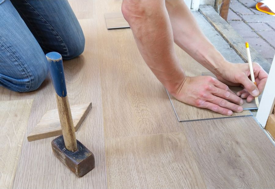 Flooring installer on his knees measuring and marking a new floor plank, with a mallet and tapping block beside him
